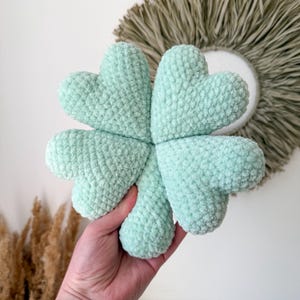 May include: A four-leaf clover shaped plush toy made of soft, mint green chenille yarn. The object features four rounded, heart-like lobes joined at the centre, creating a textured, fuzzy surface with a consistent light green colour throughout.