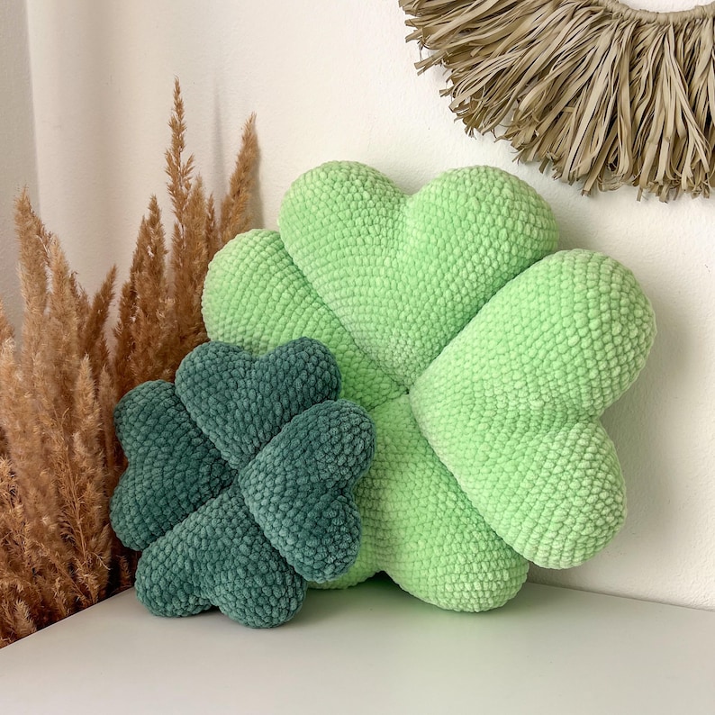 May include: Two green crocheted clover-shaped pillows. The larger pillow is light green and the smaller pillow is a darker shade of green.