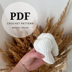 May include: A white crocheted tooth amigurumi, a handmade plush toy, held in a hand against a background of dried pampas grass. The image includes the text "PDF CROCHET PATTERN ENGLISH/POLISH".