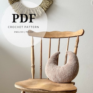 May include: PDF crochet pattern in English and Polish for a crescent-shaped pillow. The pillow is light brown and is sitting on a wooden chair.