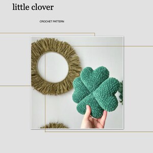 May include: A green crocheted clover-shaped pillow with a textured surface. The pillow is being held in a hand.