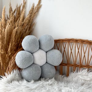 May include: A flower-shaped cushion with a central white sphere surrounded by six light blue spheres. The cushion is made of a textured, knitted material. The cushion is placed on a white faux fur rug, with a wicker chair back and dried plants in the background.