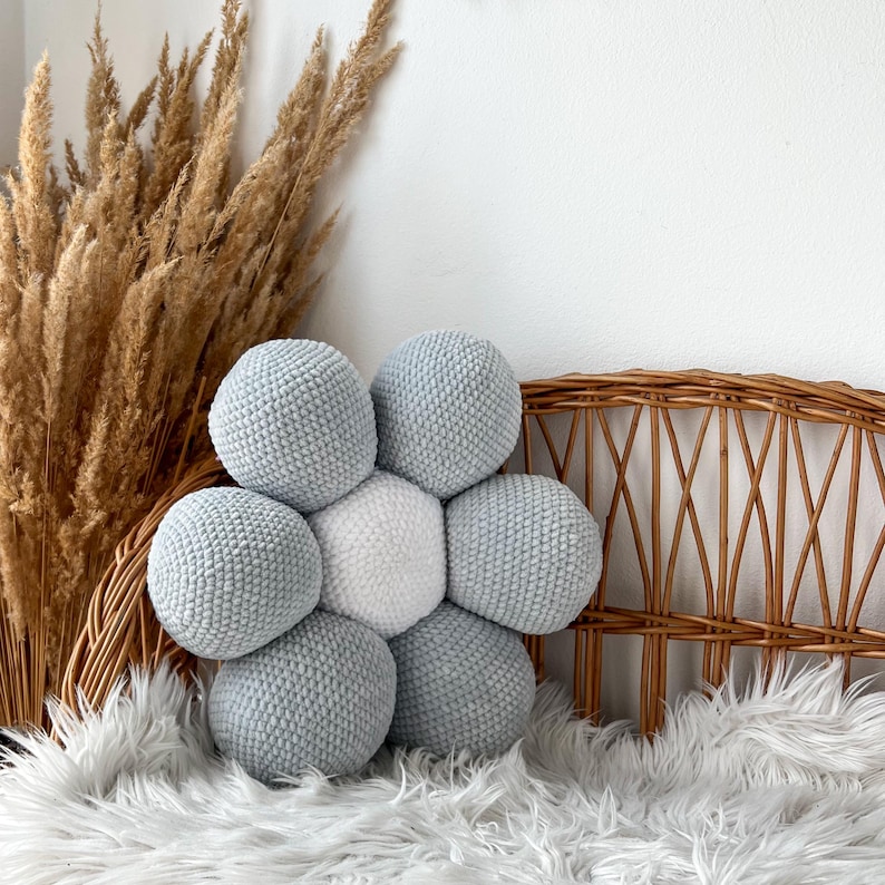 May include: A flower-shaped cushion with a central white sphere surrounded by six light blue spheres. The cushion is made of a textured, knitted material. The cushion is placed on a white faux fur rug, with a wicker chair back and dried plants in the background.