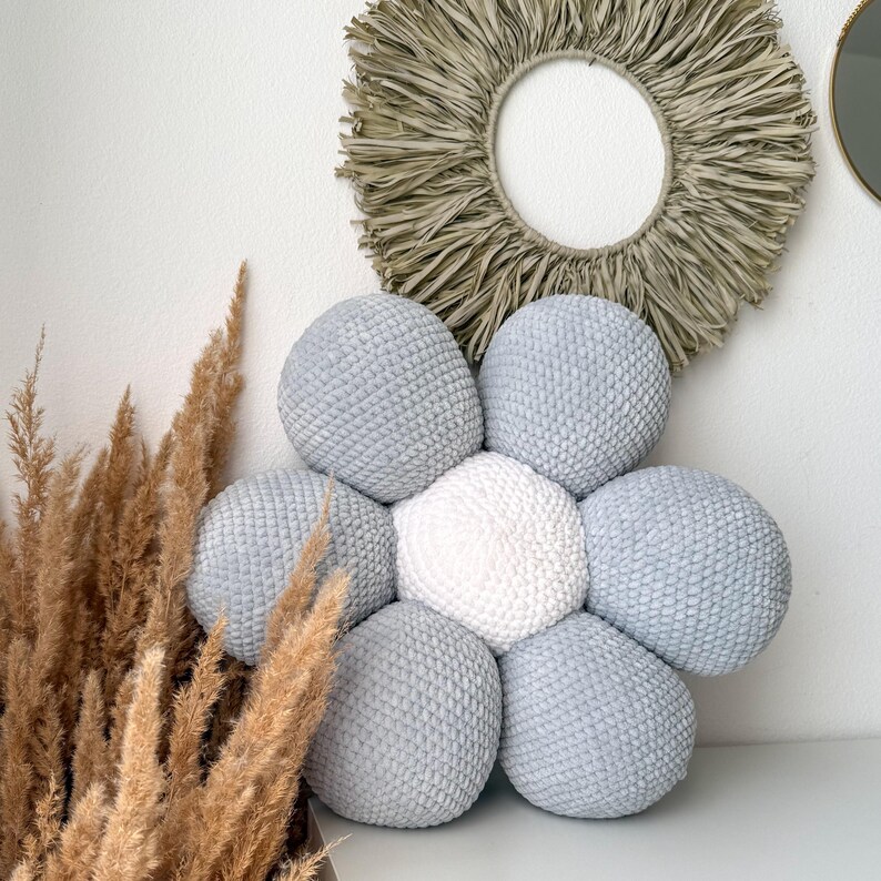 May include: A decorative, flower-shaped cushion with seven plush, round segments. Six segments are light grey, surrounding a central white segment. The cushion is on a white surface, with dried ornamental grass and a woven wall hanging.