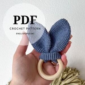 Könnte beinhalten: PDF-Häkelanleitung für eine blaue und weiße Hasenrassel mit einem Holzring. Die Anleitung ist in Englisch und Polnisch verfügbar.