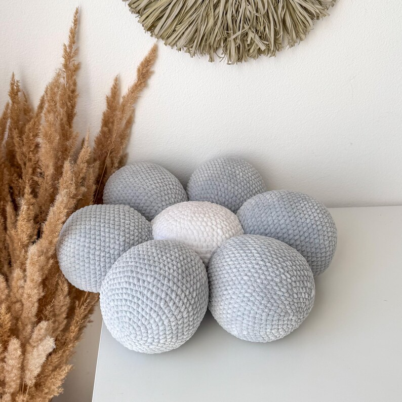 May include: A collection of seven soft, round, crocheted pillows. Six are a light blue-grey colour, arranged around a central white pillow. The pillows are on a white surface, with dried ornamental grass and a woven wall hanging in the background.