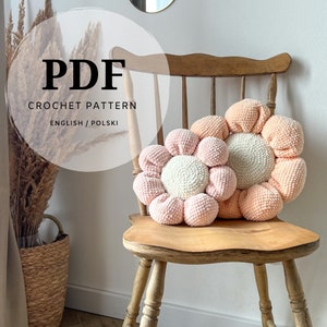 May include: Two crocheted flower-shaped cushions in pink and peach colours. The cushions are sitting on a wooden chair. The image includes the text "PDF CROCHET PATTERN ENGLISH/POLSKI".