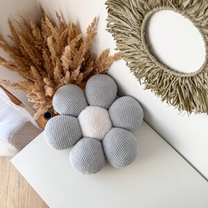 May include: A handmade, flower-shaped cushion with a central white circle and six grey petals. The cushion is made of a textured, knitted material. Dried plants and a woven wall hanging are in the background.
