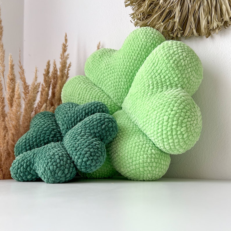May include: Two green crocheted pillows in the shape of a four-leaf clover. The larger pillow is light green and the smaller pillow is a darker green.