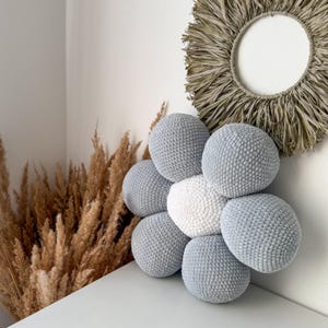 May include: A handmade, flower-shaped cushion with a central white sphere surrounded by six light grey spheres. The cushion is made of a soft, textured material. The background includes dried ornamental grass and a circular wall hanging.