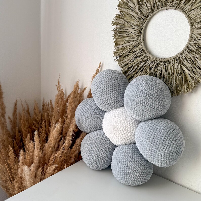 May include: A handmade, flower-shaped cushion with a central white sphere surrounded by six light grey spheres. The cushion is made of a soft, textured material. The background includes dried ornamental grass and a circular wall hanging.