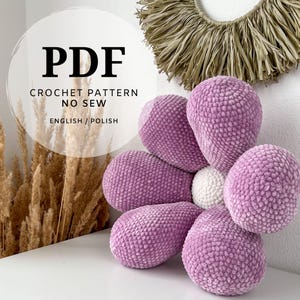 May include: PDF crochet pattern for a no-sew flower pillow. The pillow is made of soft, pink yarn and has a white center. The pattern is available in English and Polish.