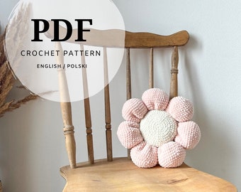 May include: A PDF crochet pattern document with text reading PDF CROCHET PATTERN ENGLISH / POLSKI, featuring a plush flower-shaped pillow with a white center and eight light pink rounded petals, resting on a wooden chair with a spindle back.