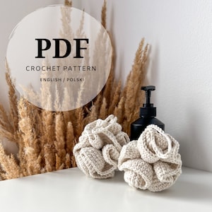 Puede incluir: Dos esponjas de baño de crochet beige sobre una superficie blanca con una botella de bomba negra y hierba de pampa seca en el fondo. El texto "PDF CROCHET PATTERN ENGLISH/POLSKI" es visible en un círculo blanco.