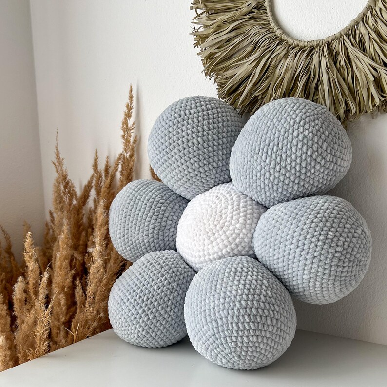 May include: A handmade, flower-shaped cushion made from seven crocheted spheres. Six are a soft grey, surrounding a central white sphere. The cushion is displayed on a white surface, with dried ornamental grasses and a woven wall hanging in the background.