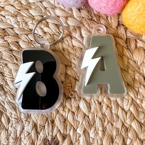 May include: Two acrylic keychains, one black with a white lightning bolt and the number 8, the other olive green with a white lightning bolt and the letter A. Both keychains have a silver keyring attached. The background is a woven mat.