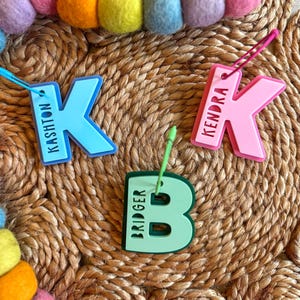May include: Three letter-shaped name tags in blue, pink, and green, spelling out the names Kashton, Kendra, and Bridger. Each tag has a hole for attaching to a bag or item. A colorful felt ball garland is in the background.