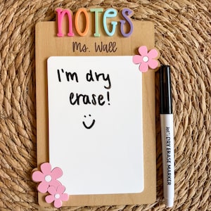 May include: A rectangular wooden dry erase board featuring the word NOTES in colorful 3D letters at the top, the text Ms. Wall, and three pink flower accents. The white board surface displays the handwritten text I'm dry erase! with a smiley face.