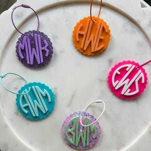 May include: A collection of personalized luggage tags in various colors, each featuring a unique monogram. The tags have a scalloped edge and a loop for attachment. Colors include purple, orange, teal, pink, and a glittery mix.