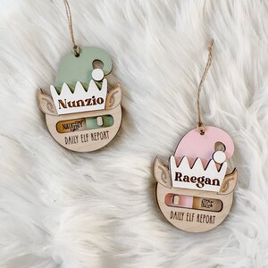 May include: Two wooden elf ornaments with a slider to indicate naughty or nice. The ornament on the left is green and says "Nunzio" and the ornament on the right is pink and says "Raegan".