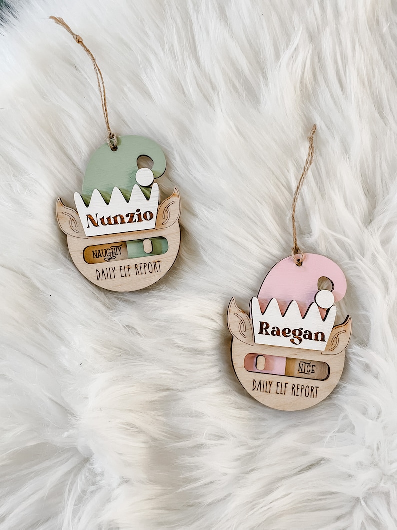 May include: Two wooden elf ornaments with a slider to indicate naughty or nice. The ornament on the left is green and says "Nunzio" and the ornament on the right is pink and says "Raegan".