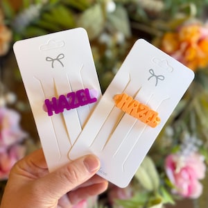 May include: Two hair clips on white cards. One clip is purple with the name "HAZEL" in bold letters. The other clip is orange with the name "HAZEL" in a heart-shaped design. Both cards have a bow design at the top.