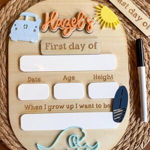 May include: A wooden milestone board featuring the text Hazel's First day of, with blank white spaces for date, age, height, and future career. It includes a light blue van, a yellow sun, a dark blue surfboard, and a light blue wave icon, plus a marker.