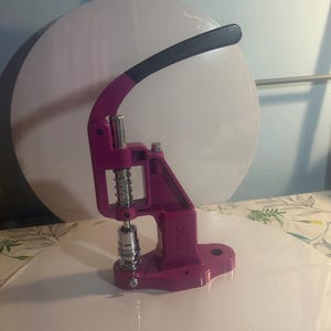 Op de afbeelding: Een magenta drukknopmachine met een zwarte handgreep. De machine heeft een veer en een zilverkleurige metalen basis. Het is ontworpen voor het aanbrengen van drukknopen op stof. De machine staat op een wit oppervlak.