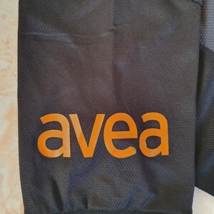 Puede incluir: Primer plano de una manga de prenda deportiva negra con la palabra "avea" en naranja impresa. La manga tiene un patr&oacute;n texturizado y una franja roja en el pu&ntilde;o.