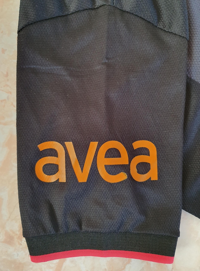 Puede incluir: Primer plano de una manga de prenda deportiva negra con la palabra "avea" en naranja impresa. La manga tiene un patr&oacute;n texturizado y una franja roja en el pu&ntilde;o.