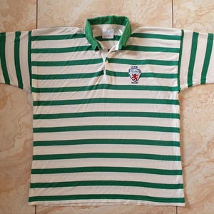 May include: A rugby shirt with horizontal green and white stripes. It has a green collar and cuffs, and a small emblem with the text "LEWES RUGBY". The shirt is displayed on a tiled surface.