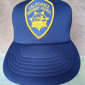 Könnte beinhalten: Eine marineblaue Trucker-Mütze mit dem Emblem "California Highway Patrol". Das Emblem ist ein gelb-blaues Schild mit dem Text "California Highway Patrol" und der Darstellung einer Person auf einem Pferd. Die Mütze hat einen gebogenen Schirm und eine Netzrückseite.