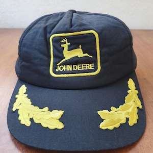 Pode incluir: Boné de beisebol azul marinho com o logotipo John Deere bordado em dourado e detalhes de folhas. O logotipo apresenta um veado. O boné tem uma viseira curva e uma coroa arredondada.