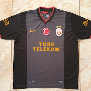 Puede incluir: Una camiseta de f&uacute;tbol del Galatasaray negra y gris con cuello en V y ribetes rojos. La camiseta presenta el logo de Nike, los emblemas del equipo y el texto "T&Uuml;RK TELEKOM" en letras doradas. Las mangas tienen letras doradas.