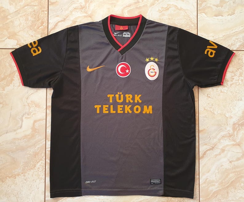 Puede incluir: Una camiseta de f&uacute;tbol del Galatasaray negra y gris con cuello en V y ribetes rojos. La camiseta presenta el logo de Nike, los emblemas del equipo y el texto "T&Uuml;RK TELEKOM" en letras doradas. Las mangas tienen letras doradas.