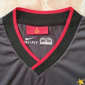 Puede incluir: Primer plano de una camiseta Nike Dri-FIT gris con cuello en V. El cuello es negro con ribete rojo. Se ve una etiqueta roja con un emblema dorado. La camiseta est&aacute; hecha en Turqu&iacute;a.