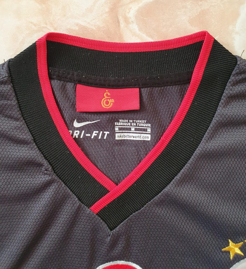 Puede incluir: Primer plano de una camiseta Nike Dri-FIT gris con cuello en V. El cuello es negro con ribete rojo. Se ve una etiqueta roja con un emblema dorado. La camiseta est&aacute; hecha en Turqu&iacute;a.