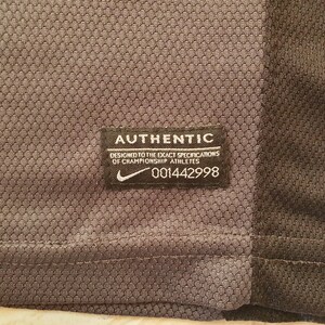 Puede incluir: Primer plano de una camiseta deportiva gris y negra. Una etiqueta rectangular negra con texto blanco dice "AUTHENTIC" y "DESIGNED TO THE EXACT SPECIFICATIONS OF CHAMPIONSHIP ATHLETES." El logo de Nike es visible.