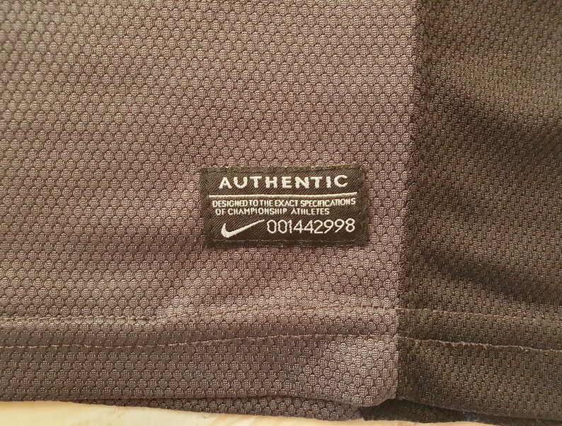 Puede incluir: Primer plano de una camiseta deportiva gris y negra. Una etiqueta rectangular negra con texto blanco dice "AUTHENTIC" y "DESIGNED TO THE EXACT SPECIFICATIONS OF CHAMPIONSHIP ATHLETES." El logo de Nike es visible.