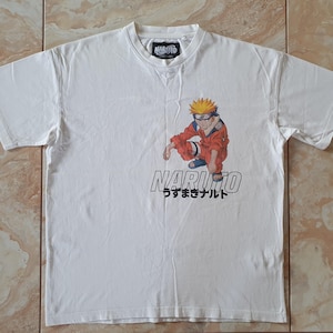 May include: White t-shirt with a graphic of Naruto, an anime character, in an orange and blue outfit. The word "NARUTO" is printed in black, with Japanese characters below. The shirt has a crew neck and short sleeves.