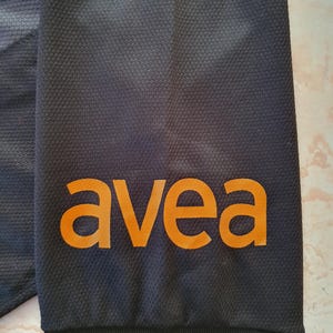 Puede incluir: Primer plano de una manga de camiseta deportiva negra con la palabra naranja "avea" impresa. La manga tiene un patr&oacute;n texturizado y una raya roja en el pu&ntilde;o. Un logotipo del equipo es visible en la esquina superior izquierda.