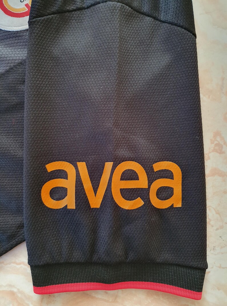 Puede incluir: Primer plano de una manga de camiseta deportiva negra con la palabra naranja "avea" impresa. La manga tiene un patr&oacute;n texturizado y una raya roja en el pu&ntilde;o. Un logotipo del equipo es visible en la esquina superior izquierda.