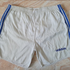 May include: White athletic shorts with blue stripes down the sides and a drawstring waist. The Adidas logo is embroidered in blue on the left leg. These shorts are made of a lightweight material.