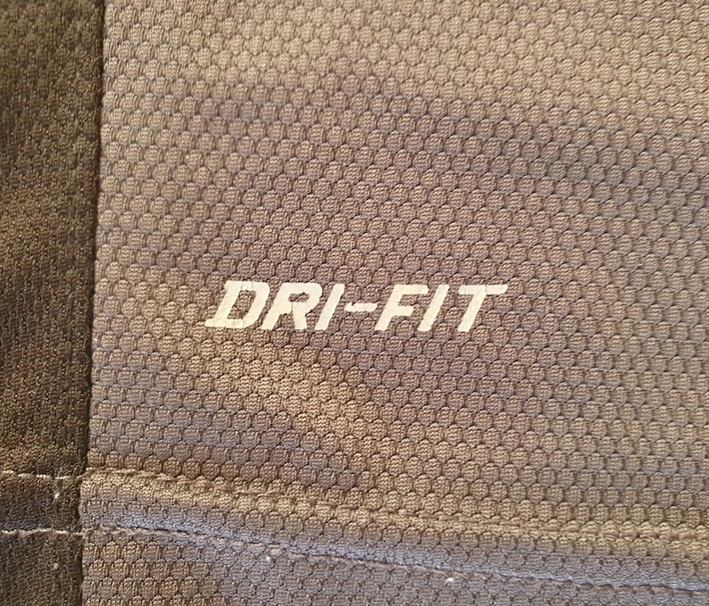 Puede incluir: Primer plano de una camiseta deportiva gris con el texto "DRI-FIT" en blanco. La camiseta tiene un patr&oacute;n texturizado en forma de panal. Un panel gris m&aacute;s oscuro es visible en el lado izquierdo de la imagen.