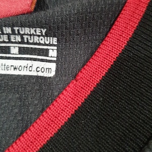 Puede incluir: Primer plano de una camiseta deportiva gris oscuro con una raya roja a lo largo del cuello y el hombro. La camiseta tiene una etiqueta con el texto "MADE IN TURKEY" y "betterworld.com". La camiseta es talla M.