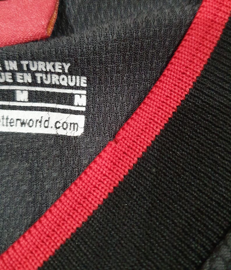 Puede incluir: Primer plano de una camiseta deportiva gris oscuro con una raya roja a lo largo del cuello y el hombro. La camiseta tiene una etiqueta con el texto "MADE IN TURKEY" y "betterworld.com". La camiseta es talla M.