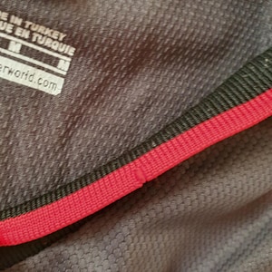 Puede incluir: Primer plano de una camiseta deportiva gris con un patr&oacute;n texturizado. La camiseta tiene un ribete rojo y negro alrededor del cuello. La etiqueta dice "MADE IN TURKEY" y "M". La parte inferior de la camiseta tiene dos estrellas amarillas.