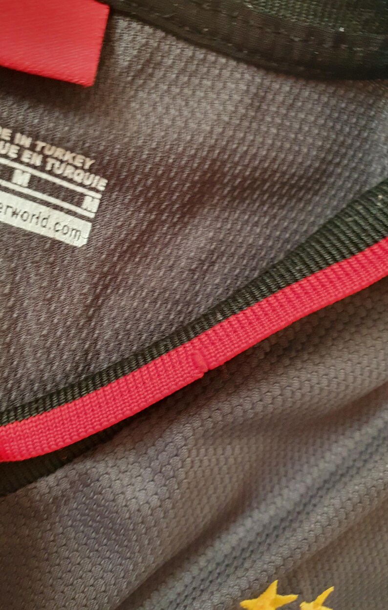 Puede incluir: Primer plano de una camiseta deportiva gris con un patr&oacute;n texturizado. La camiseta tiene un ribete rojo y negro alrededor del cuello. La etiqueta dice "MADE IN TURKEY" y "M". La parte inferior de la camiseta tiene dos estrellas amarillas.