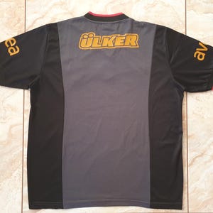 Puede incluir: Camiseta deportiva negra y gris con la palabra "&Uuml;LKER" en letras doradas en la parte superior de la espalda. Las mangas tienen letras doradas y ribetes rojos. La camiseta tiene un panel central gris.