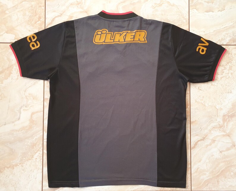 Puede incluir: Camiseta deportiva negra y gris con la palabra "&Uuml;LKER" en letras doradas en la parte superior de la espalda. Las mangas tienen letras doradas y ribetes rojos. La camiseta tiene un panel central gris.